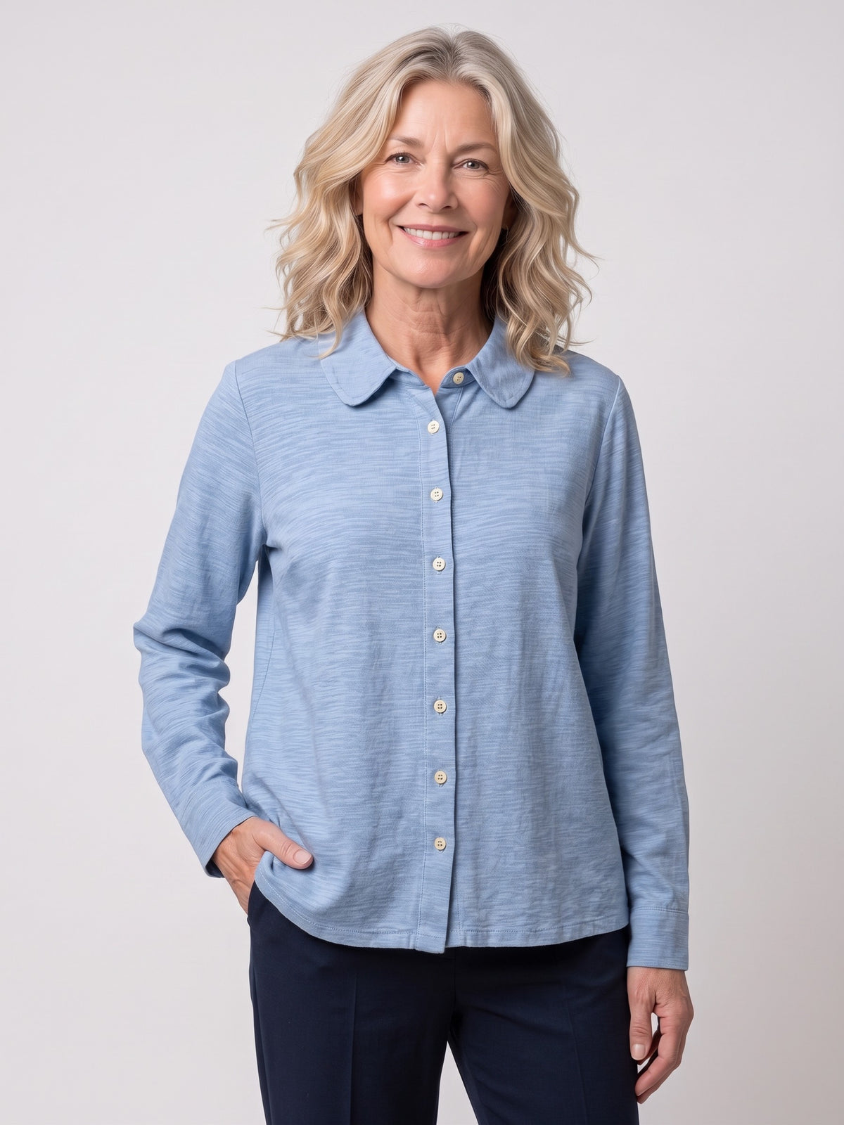 A smiling blonde woman wearing a light blue long-sleeve Fitted button-up shirt in Linen Cotton with a collar and dark navy pants, standing against a light gray studio background.
