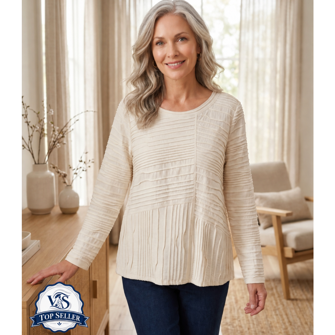 A smiling woman with gray hair stands indoors wearing a cream ribbed long-sleeve top and dark blue jeans. The room has soft beige curtains, a wooden cabinet, and a light upholstered chair in the background.