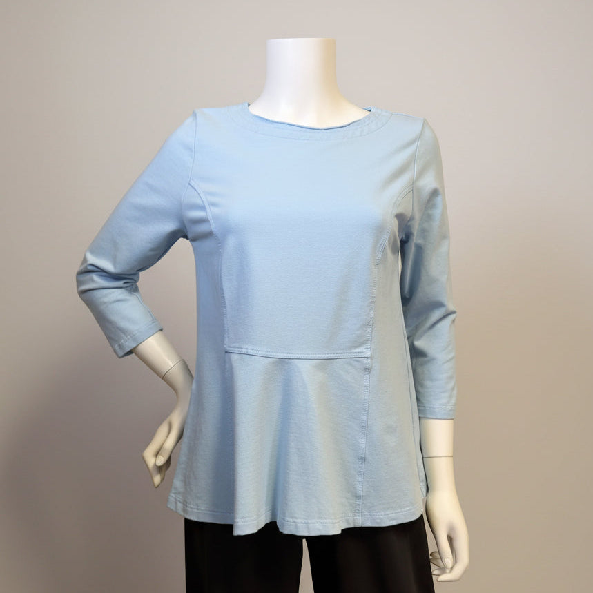 A light blue long-sleeve top displayed on a mannequin, paired with black pants, set against a beige fabric backdrop.