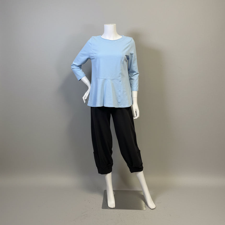A light blue long-sleeve peplum top displayed on a mannequin with black cropped pants, set against a beige fabric backdrop with a wooden cabinet on the left.