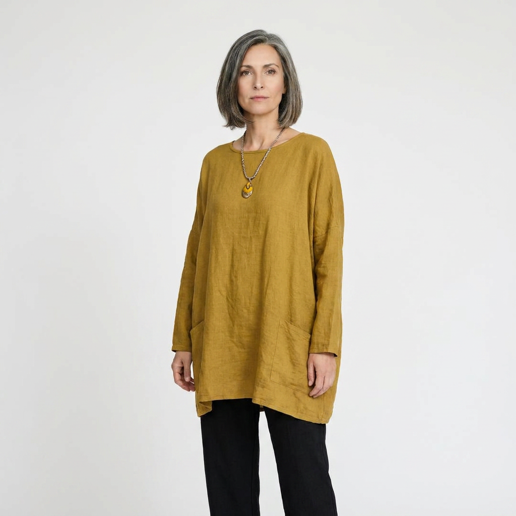 A woman with short gray hair wearing a loose, mustard yellow linen tunic top and black pants, standing against a plain light gray studio background.