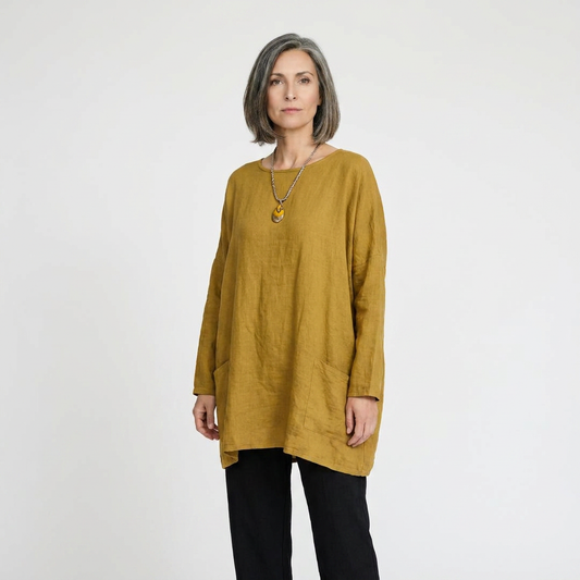 A woman with short gray hair wearing a loose, mustard yellow linen tunic top and black pants, standing against a plain light gray studio background.